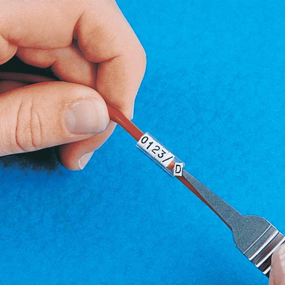 A hand slides a small white element marked 0 into a sleeve on a red wire using a metal tool, against a blue background — showcasing the precision of Grafoplast® labelling solutions.