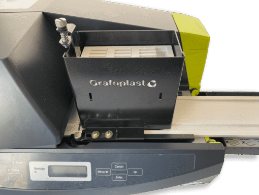 Close-up of a Grafoplast® EVOMAX thermal printer with its black metal cartridge holder open, displaying white label strips inside. Control buttons and a small display are visible on the lower front panel.