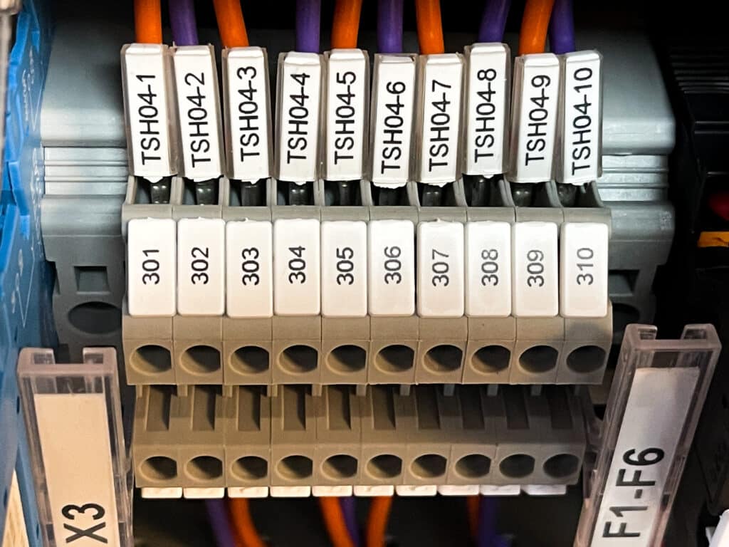 Close-up of a terminal block with labelled connections and wires, illustrating best practices for marking electrical panels and components. Labels include TSH04-1 to TSH04-10 above, numbers 301 to 310 below, with orange and purple wires attached.