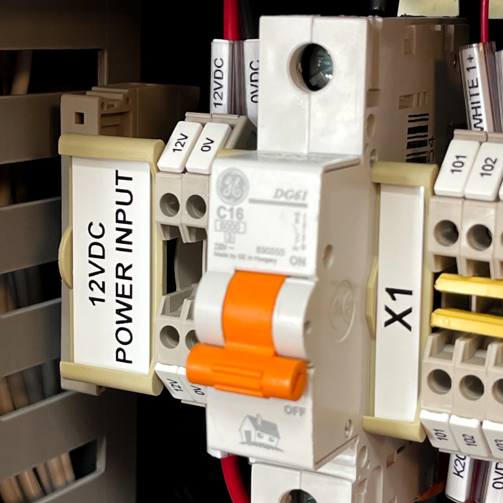Close-up of an electrical panel featuring a 12VDC power input label, orange circuit breaker switch, terminal block marking, and various labelled wiring terminals with connected red, black, and white wires.